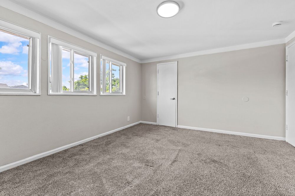 Spacious empty bedroom with gray carpet, crown molding, large windows, and a white door in a modern home.