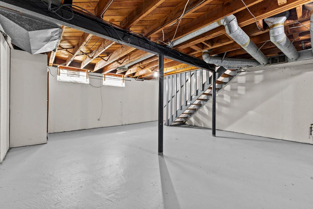 Unfinished basement with painted concrete floors, exposed wood joists, white walls, and open staircase.