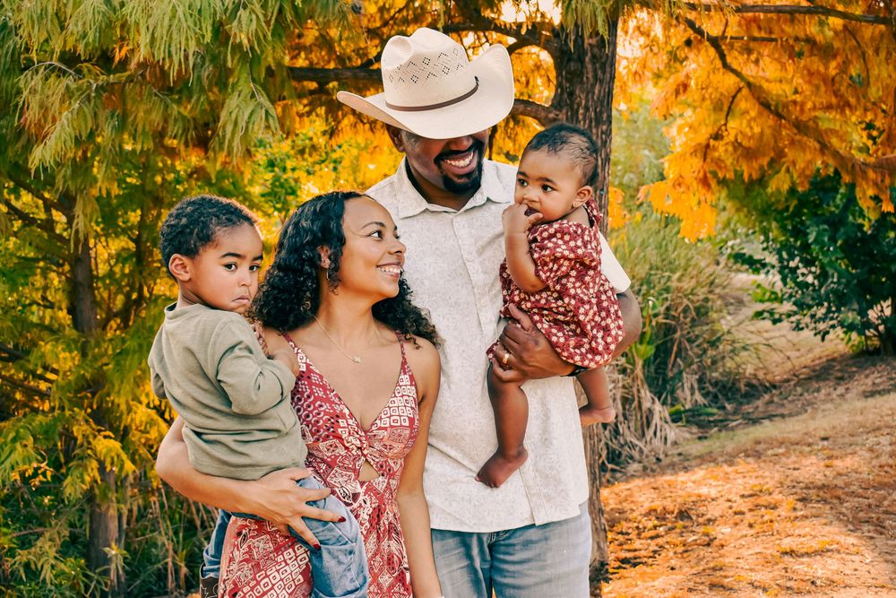 A happy Black family of four poses outdoors among golden autumn trees, parents holding two young children.