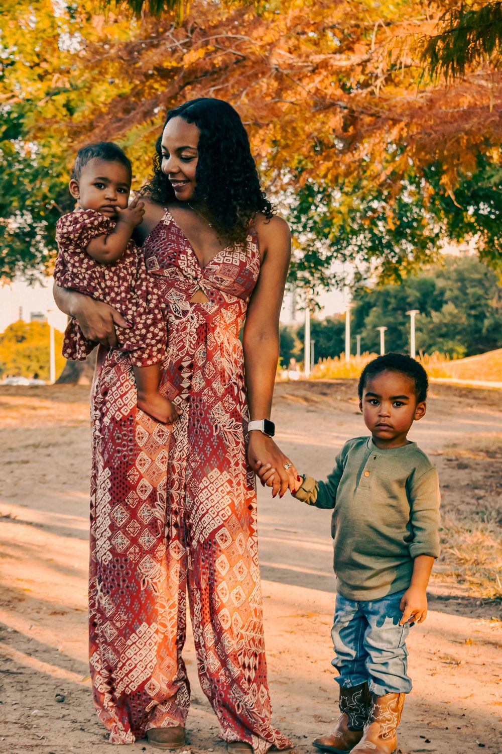A mother in a red patterned jumpsuit holds her baby while holding hands with her young son outdoors in autumn.