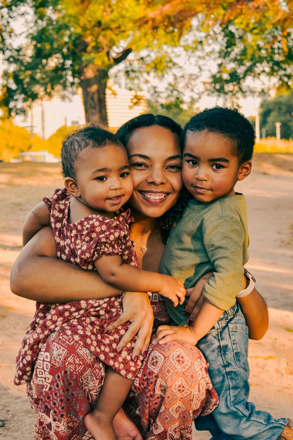 A smiling woman hugs two young children outdoors near a tree during golden hour.