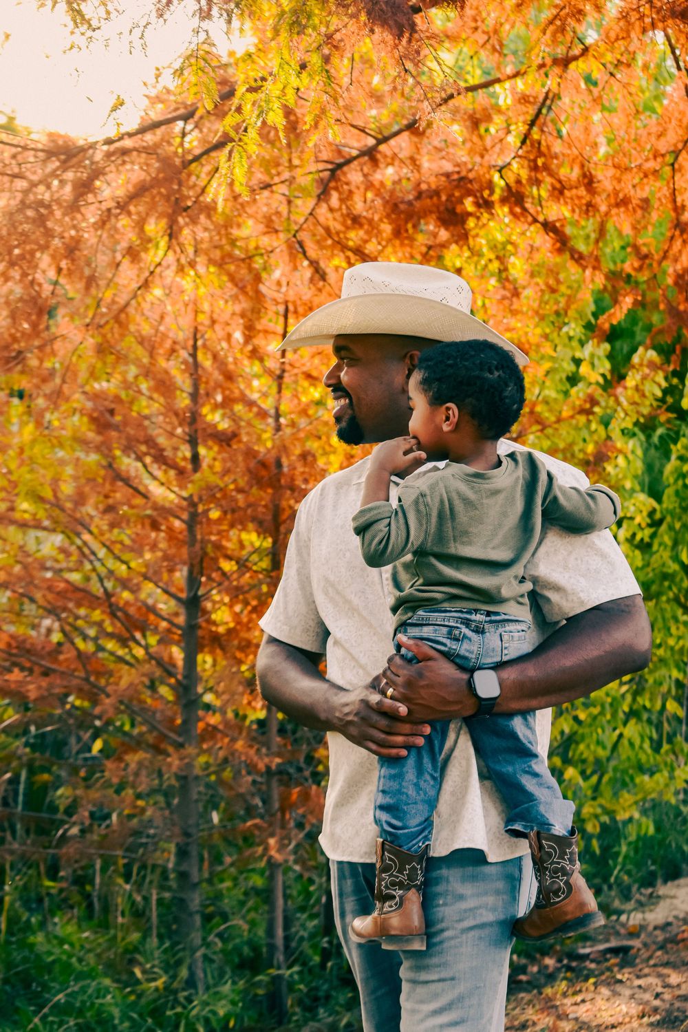 A laughing father in a cowboy hat holds his young son among vibrant orange and yellow autumn trees.