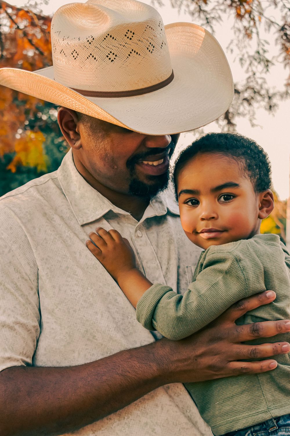 A smiling Black man wearing a straw cowboy hat holds a young toddler boy outdoors during autumn.