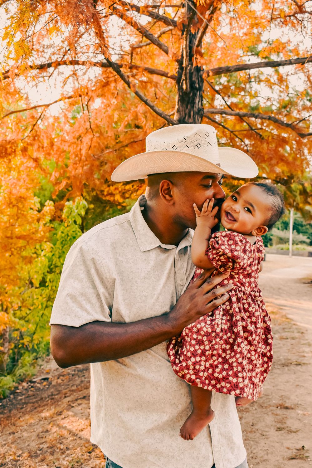 Father in cowboy hat kissing his baby daughter in red floral dress among vibrant autumn foliage.