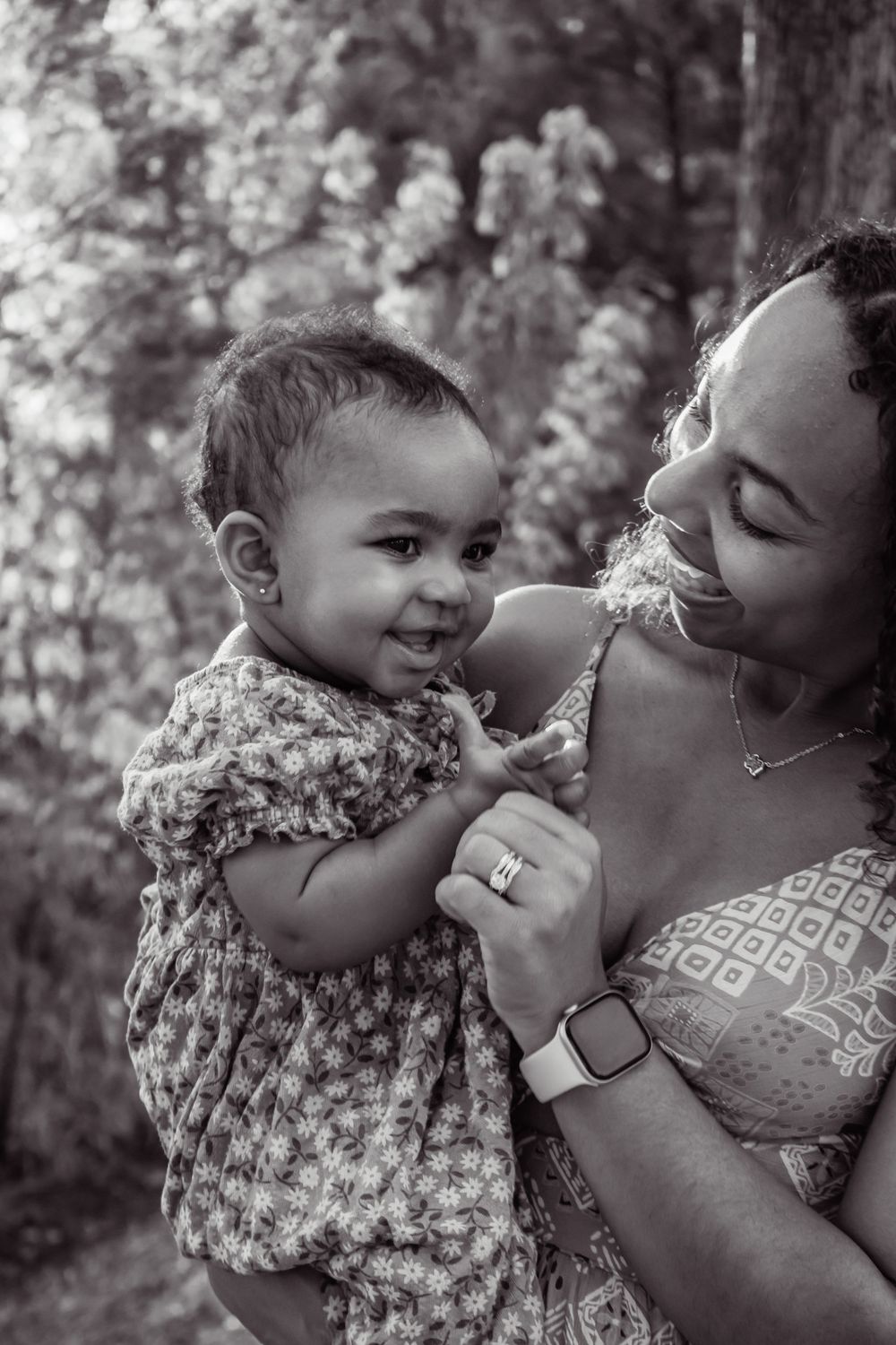 Black and white photo of a smiling mother holding her happy baby girl outdoors near blooming trees.