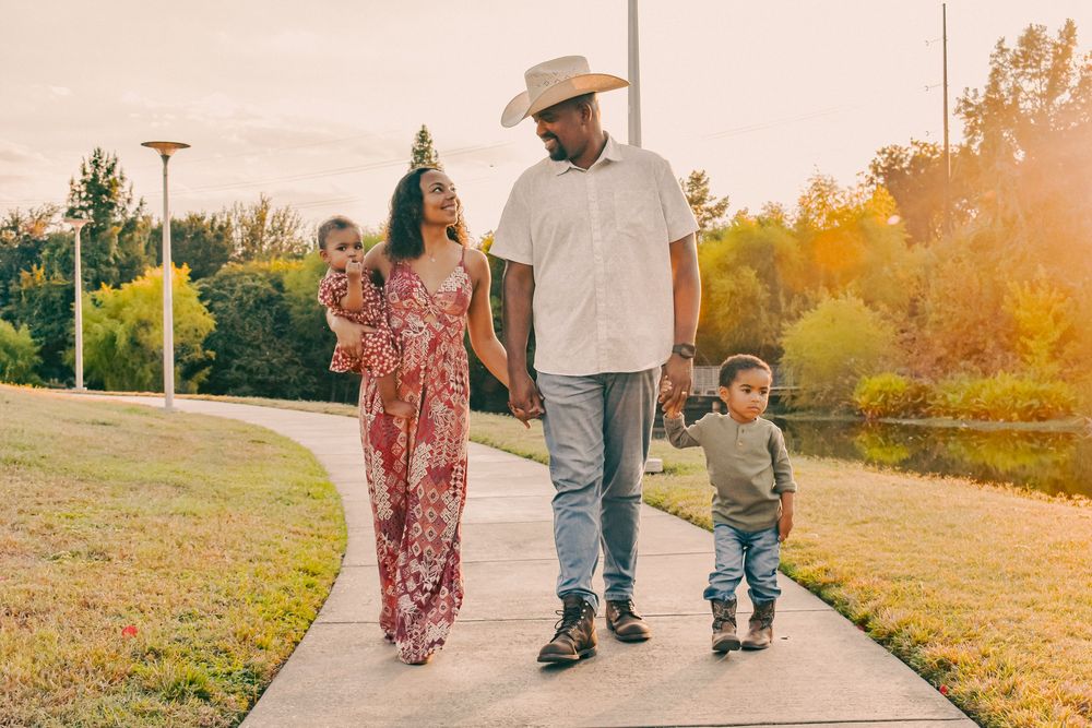 Family of four walking on a park path at sunset, parents holding children's hands in a lush green setting.