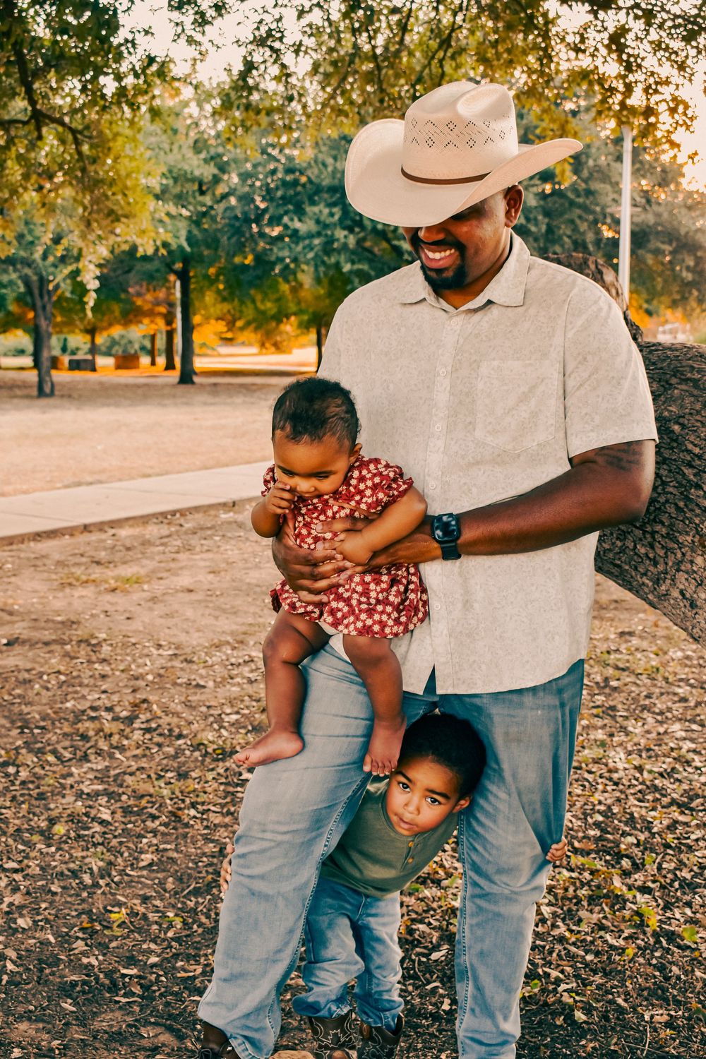 A smiling Black father in a cowboy hat holds his baby girl while his young son peeks out from behind him in a park.