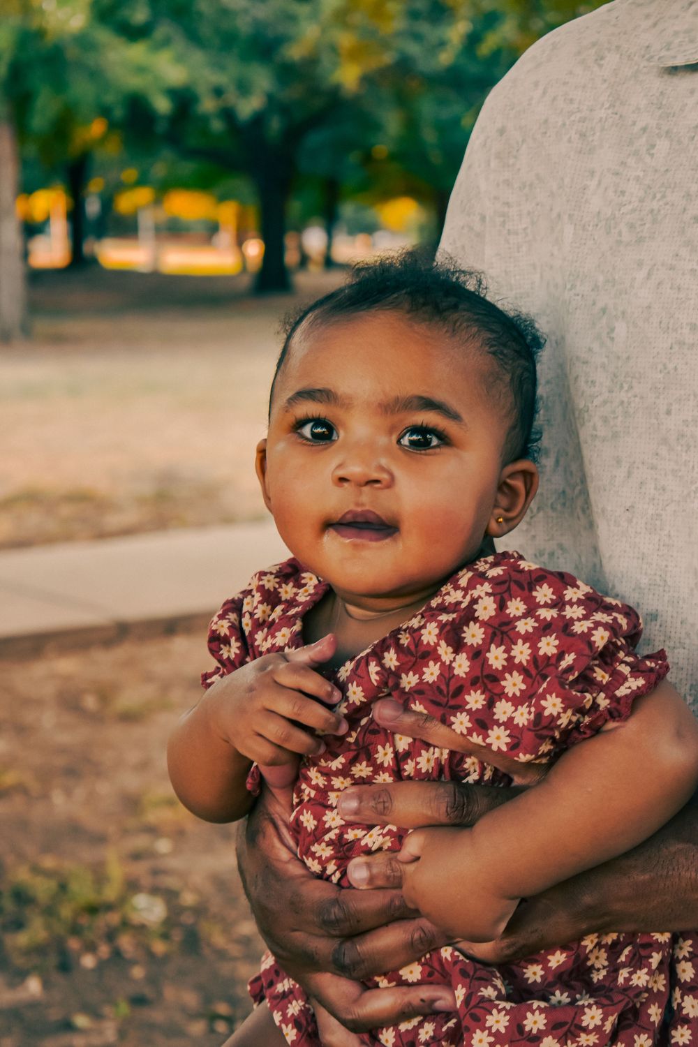 Adorable baby girl in a red floral dress being held outdoors in a park during golden hour.