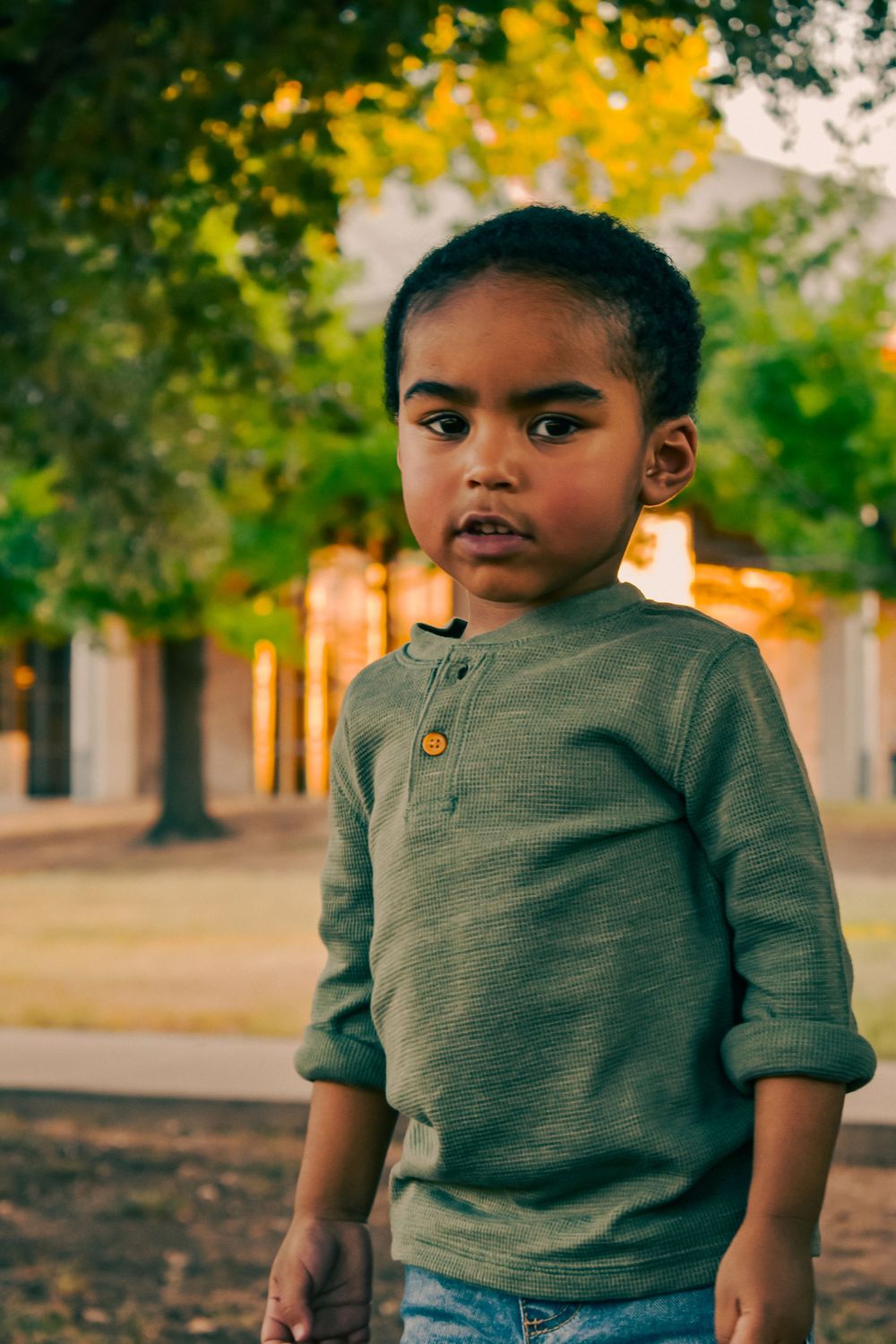 Young Black boy in green henley shirt stands outdoors near trees during golden hour, looking at camera.