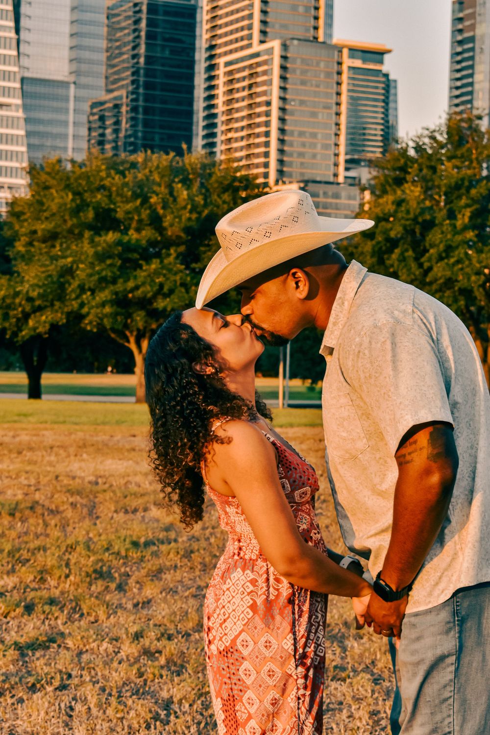 Couple sharing a romantic kiss in a park with city skyline backdrop during golden hour sunset.