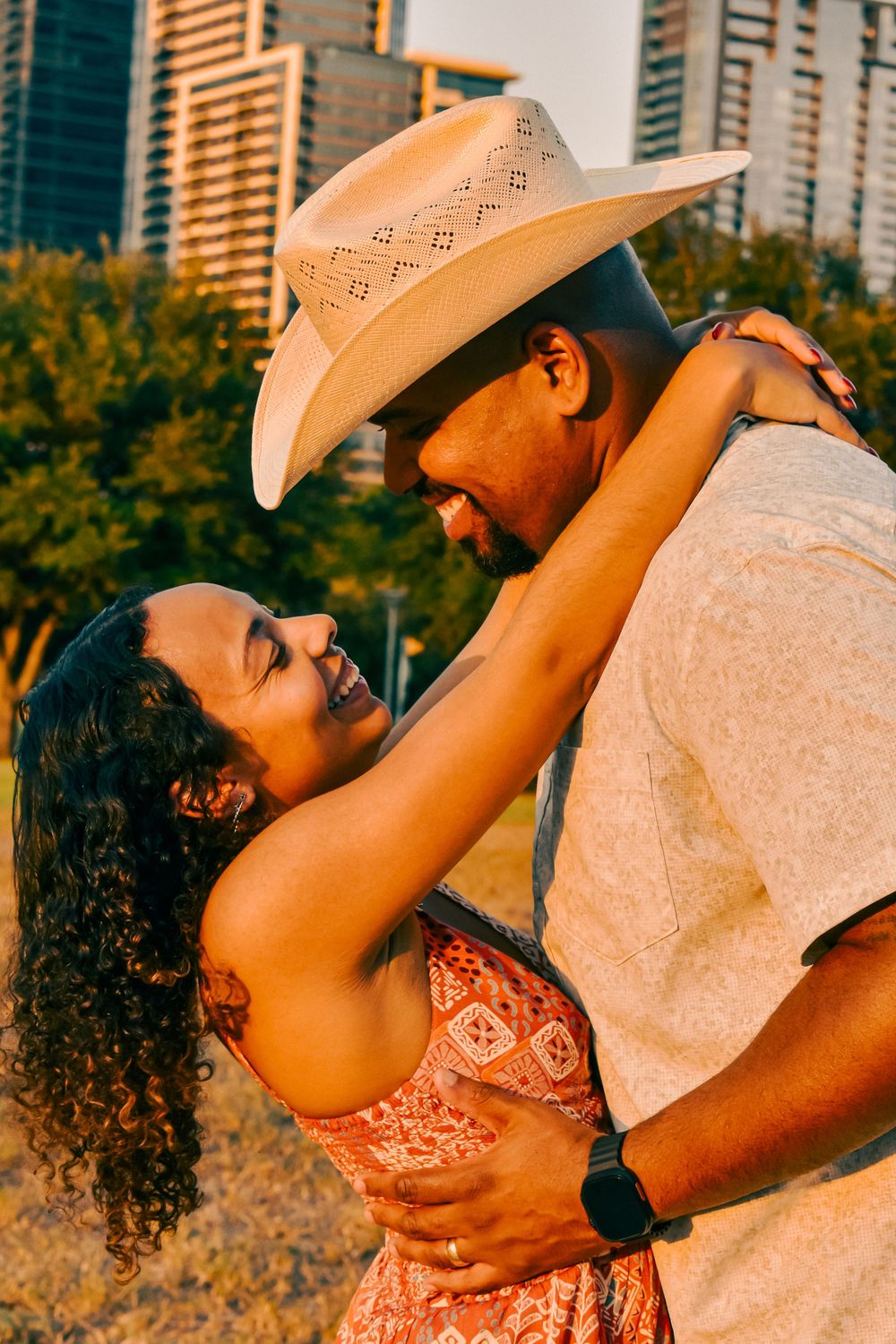 A couple embraces outdoors, man in cowboy hat and woman in floral dress, smiling at each other with city skyline behind.