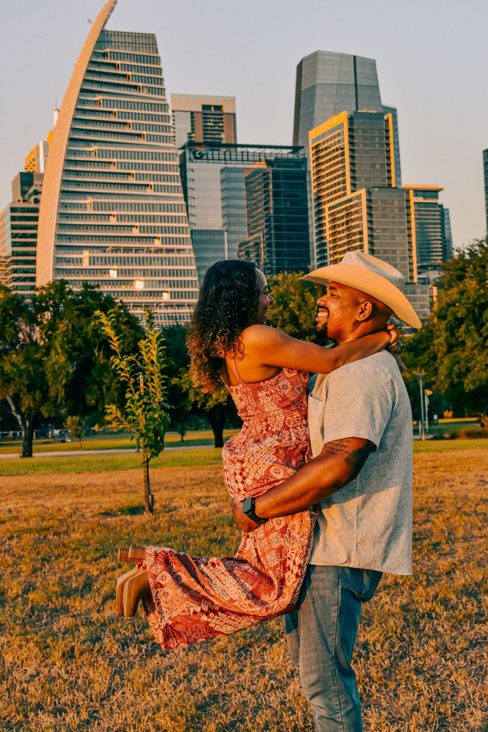 A joyful couple embraces in a park with the Dallas skyline at golden hour, man in cowboy hat lifting smiling woman.