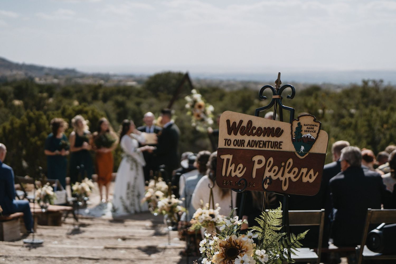 Outdoor wedding ceremony with a wooden welcome sign reading The Pfeifers adorned with sunflowers at Elena Gallegos Trailhead