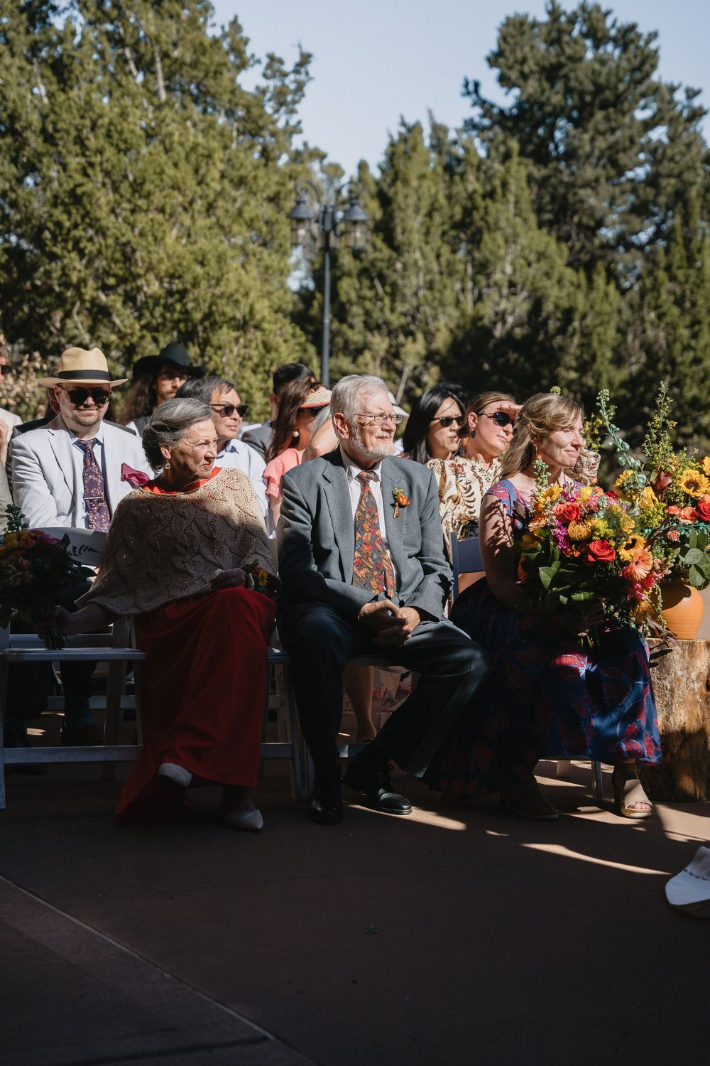 Wedding guests seated outdoors enjoying ceremony with colorful floral arrangements nearby.