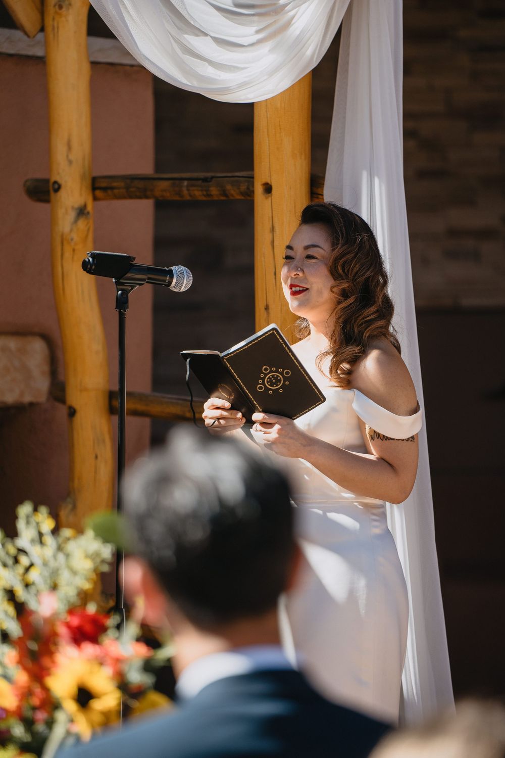 Bride smiles while reading vows from book at decorated outdoor wedding ceremony altar.