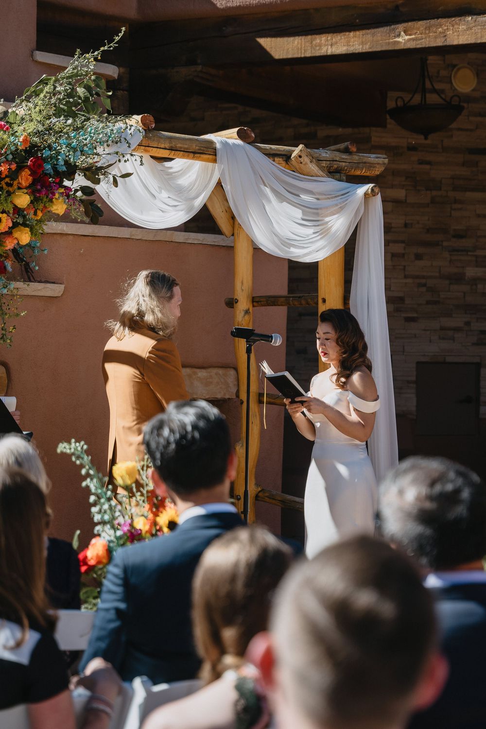 Bride reads vows at rustic wooden arch altar with white draping and floral decor.
