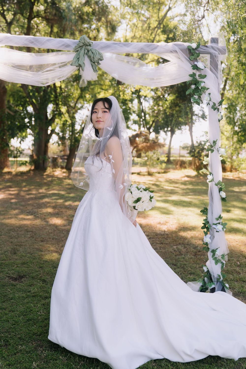Elegant bride in white ball gown and veil holding bouquet under floral arch outdoors.
