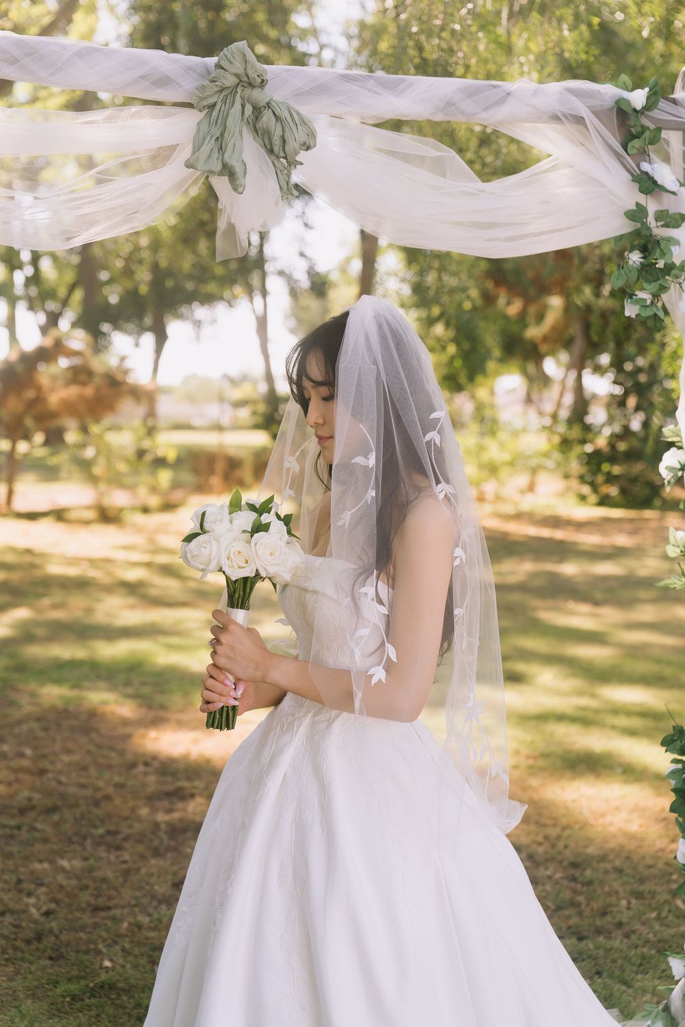 Bride in white gown and veil holds white rose bouquet outdoors under decorated arch.