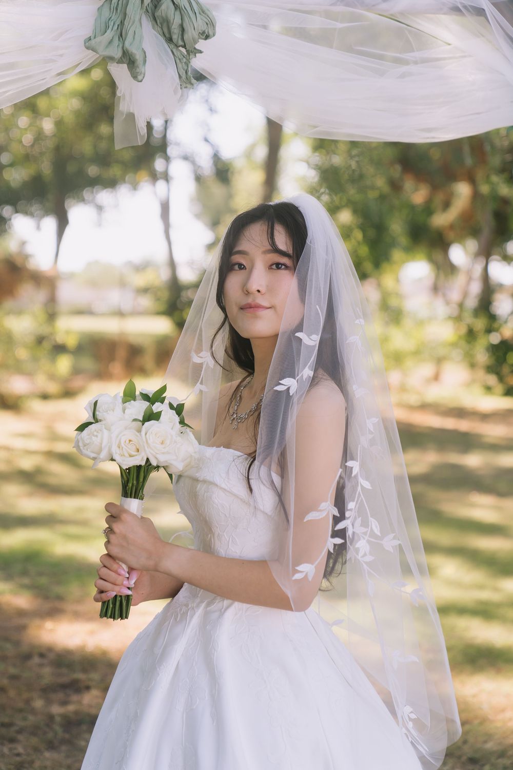 Beautiful Asian bride in white dress and veil holding white rose bouquet outdoors.
