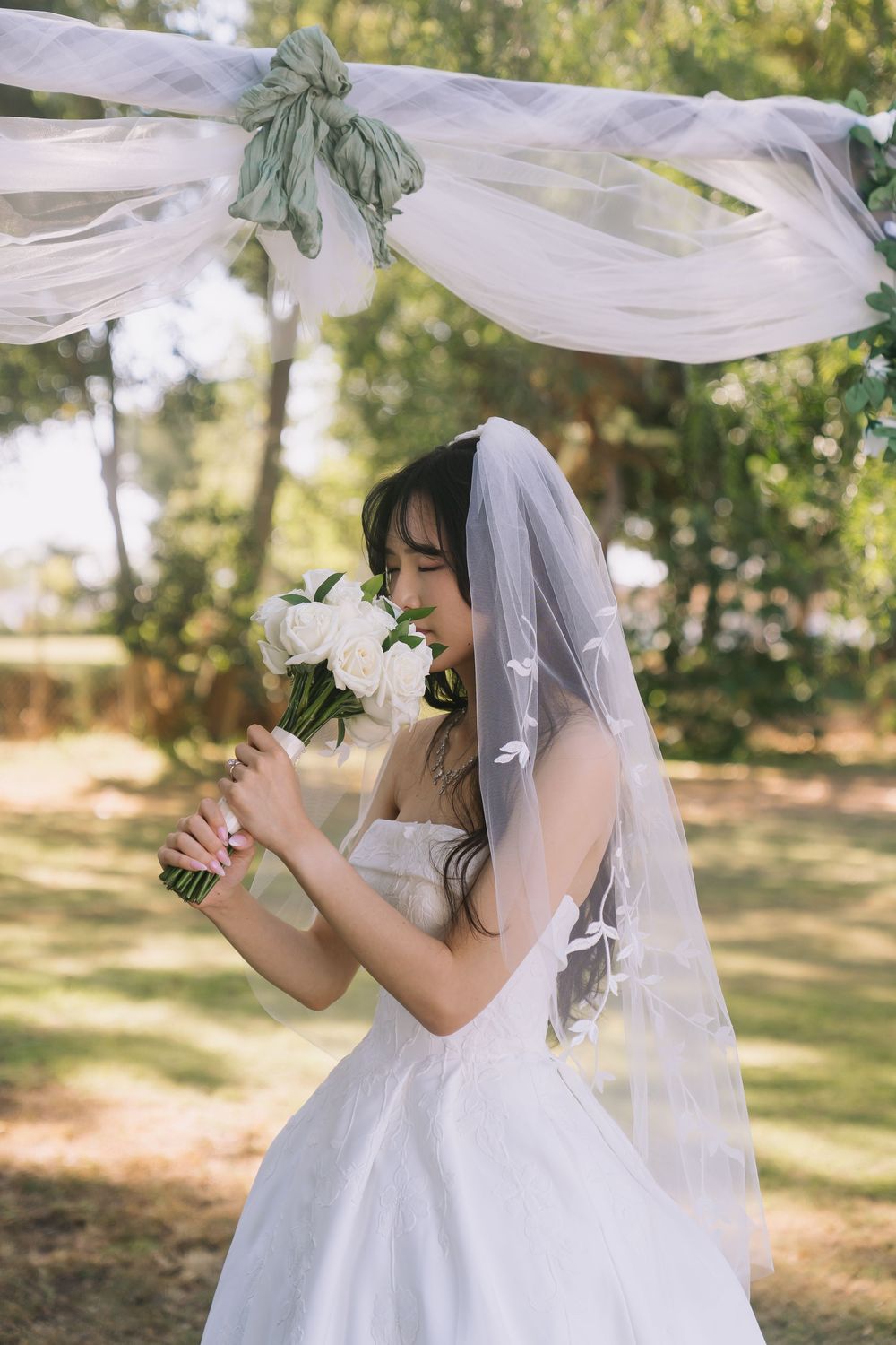 Bride in white strapless gown and veil smelling white flower bouquet under decorated arch in sunny garden.