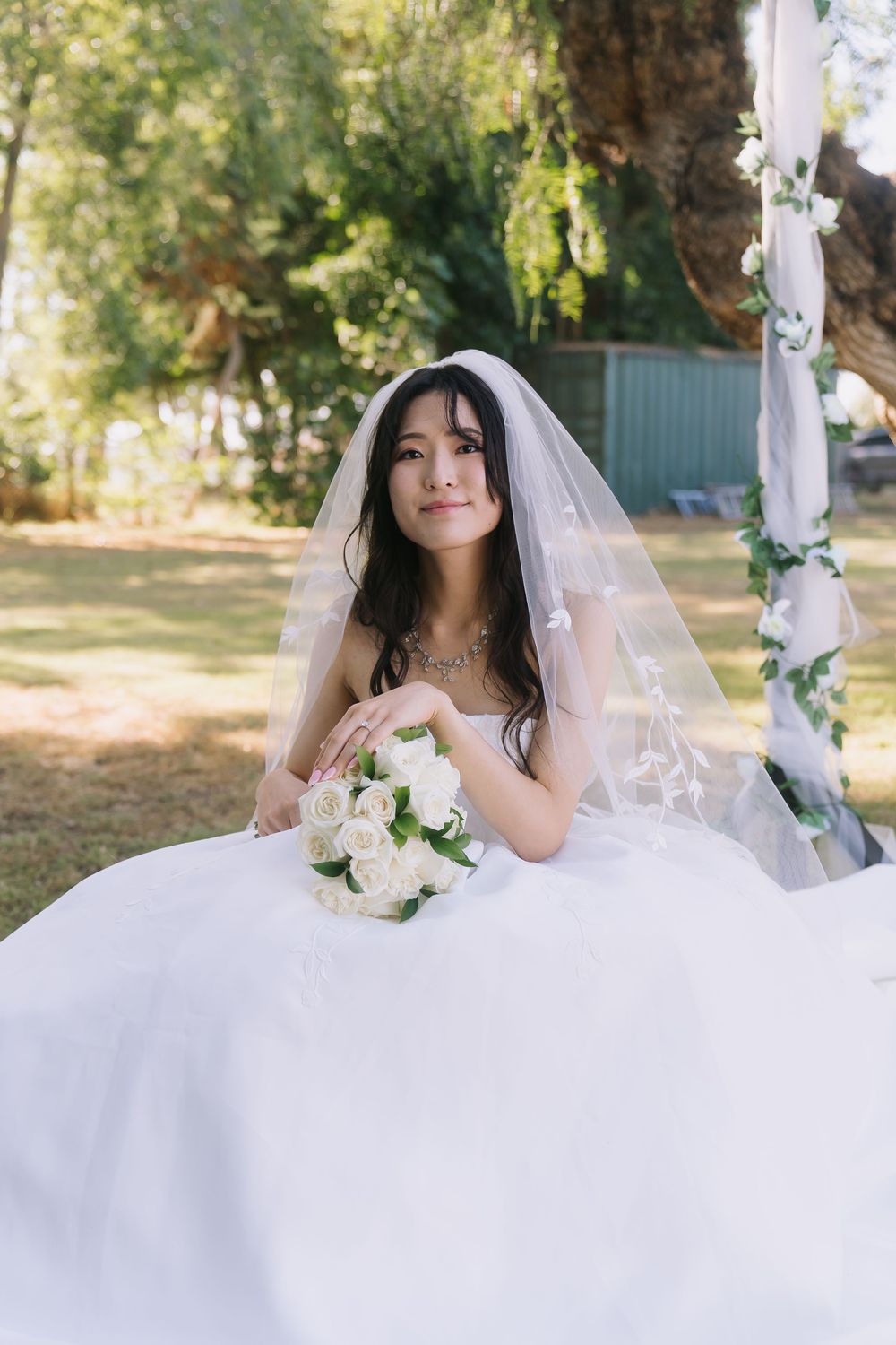 Elegant bride in white dress and veil sits outdoors holding white rose bouquet.