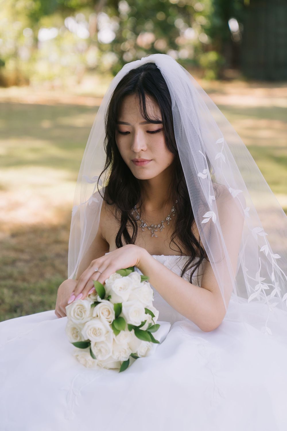 Bride in white gown and veil looks down at bouquet of white roses on table in outdoor garden setting.