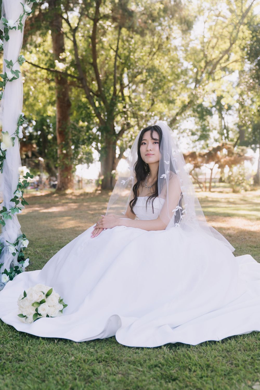 Elegant bride in full white ball gown and veil sits on green grass beneath shady trees outdoors.