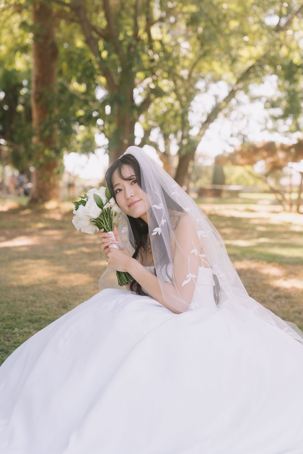 Beautiful bride in white wedding dress and veil holds white flower bouquet while sitting on grass.