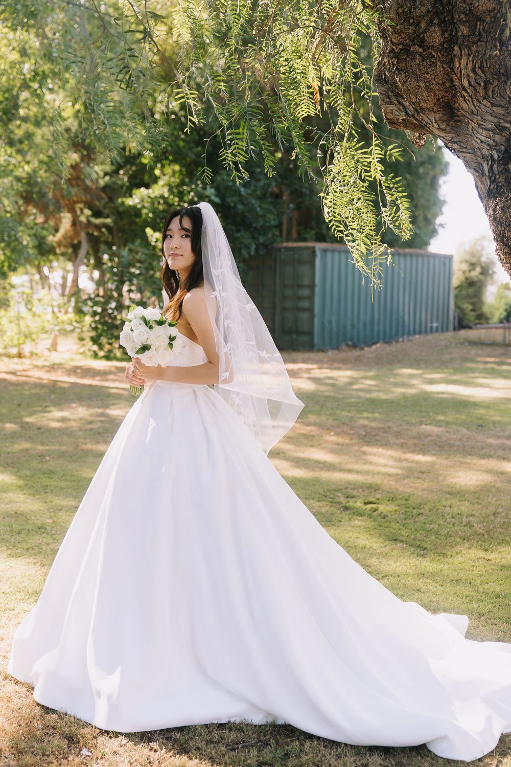 Bride in white ball gown with veil holds bouquet outdoors under green tree on sunny wedding day.