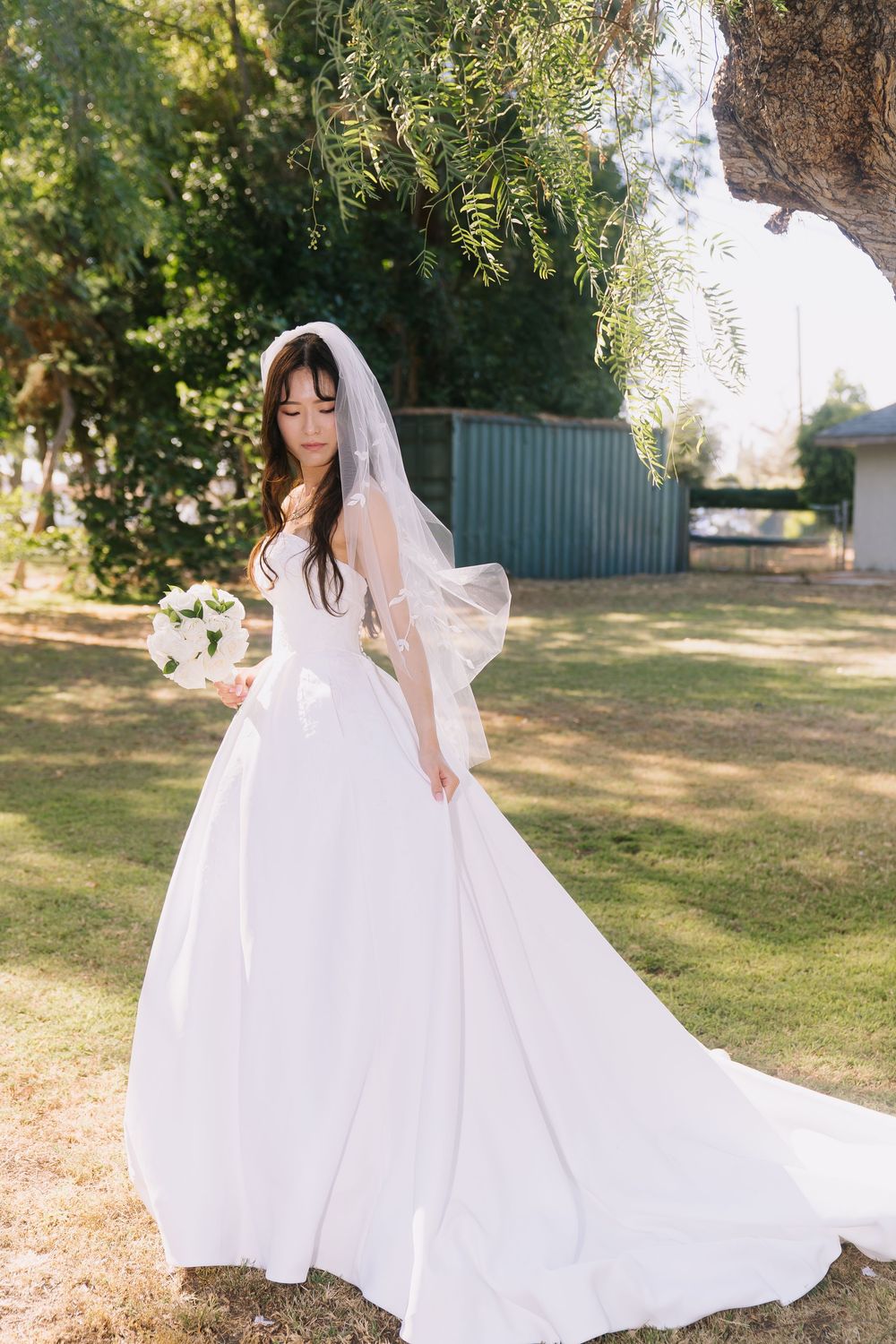 Bride in elegant white ball gown with veil holding bouquet standing under tree in sunlit garden.