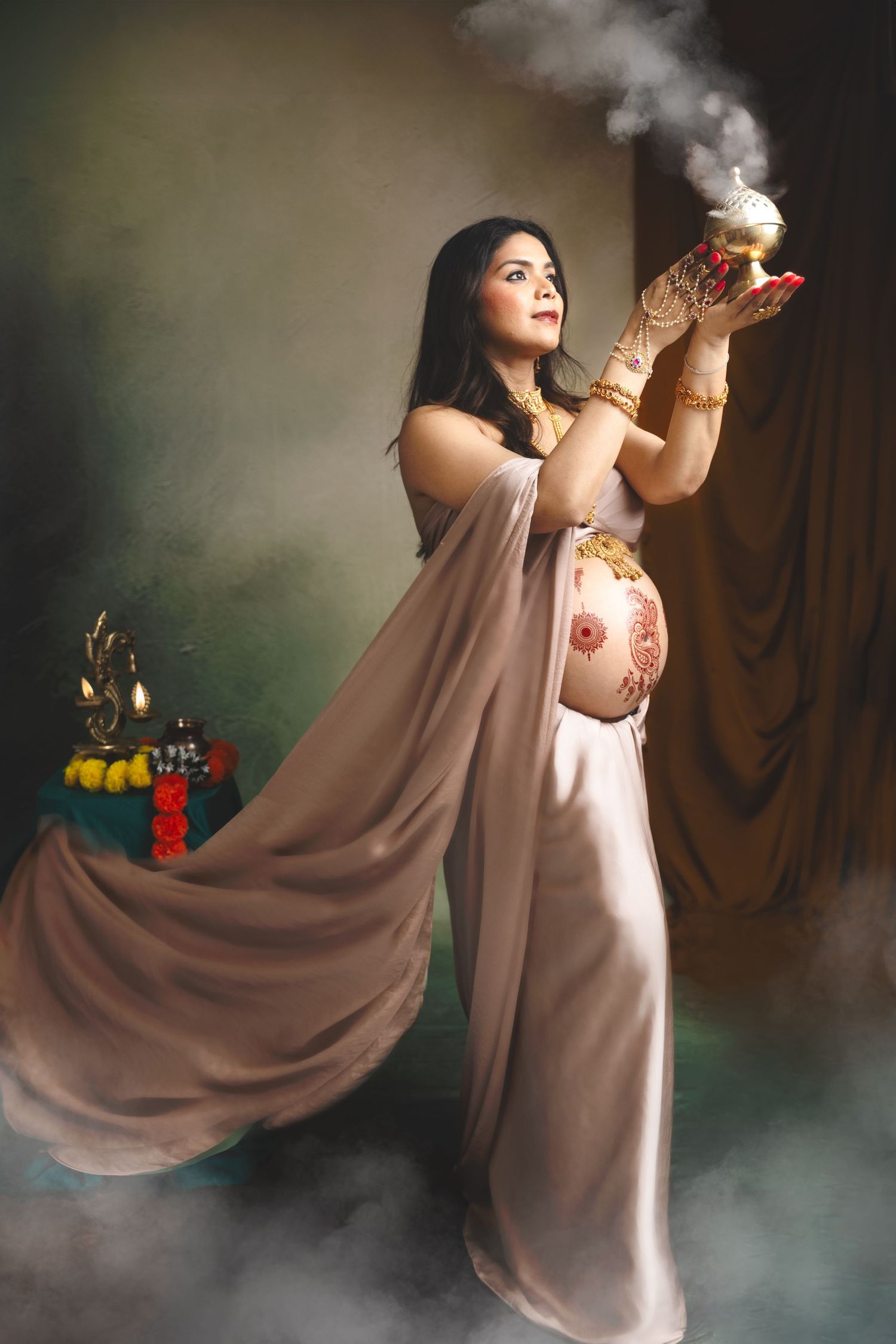 Pregnant Indian woman in elegant drape holding smoking diya, adorned with mehndi and gold jewelry in studio setting.