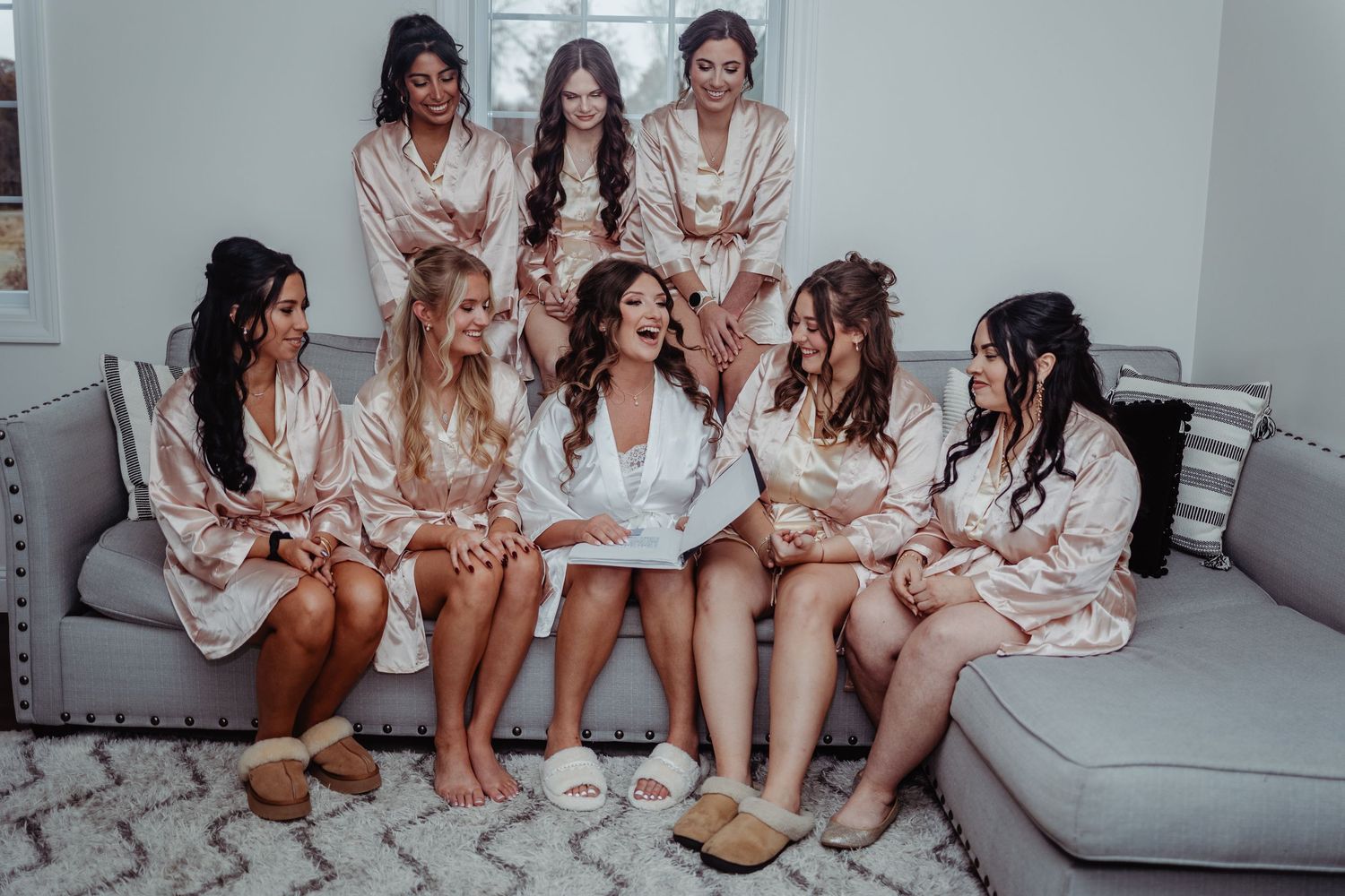 A bride in white robe laughs with seven bridesmaids in matching satin robes seated on a gray sofa.