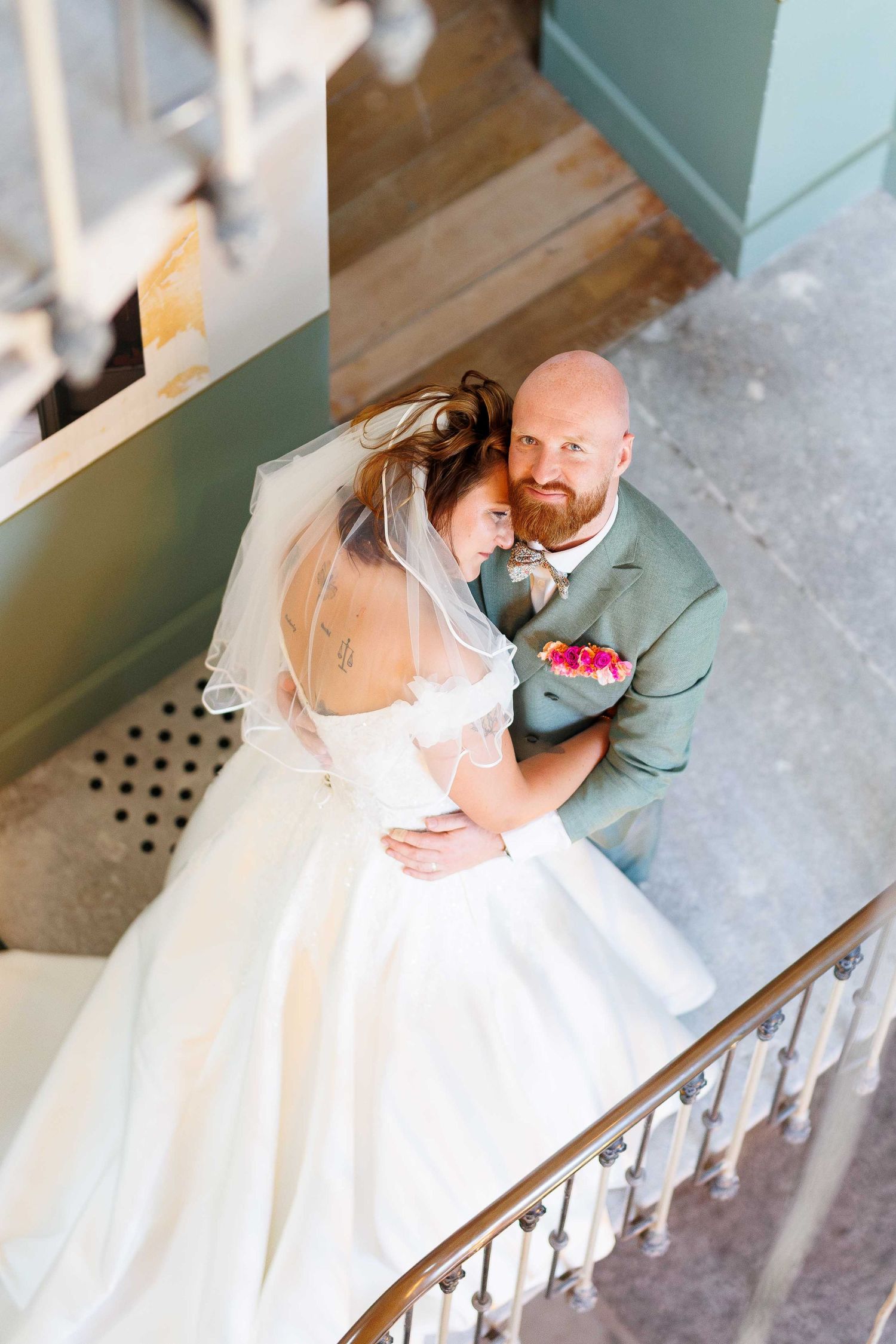 Aerial view of bride and groom embracing on staircase, bride in white gown with veil, groom in sage suit holding bouquet.