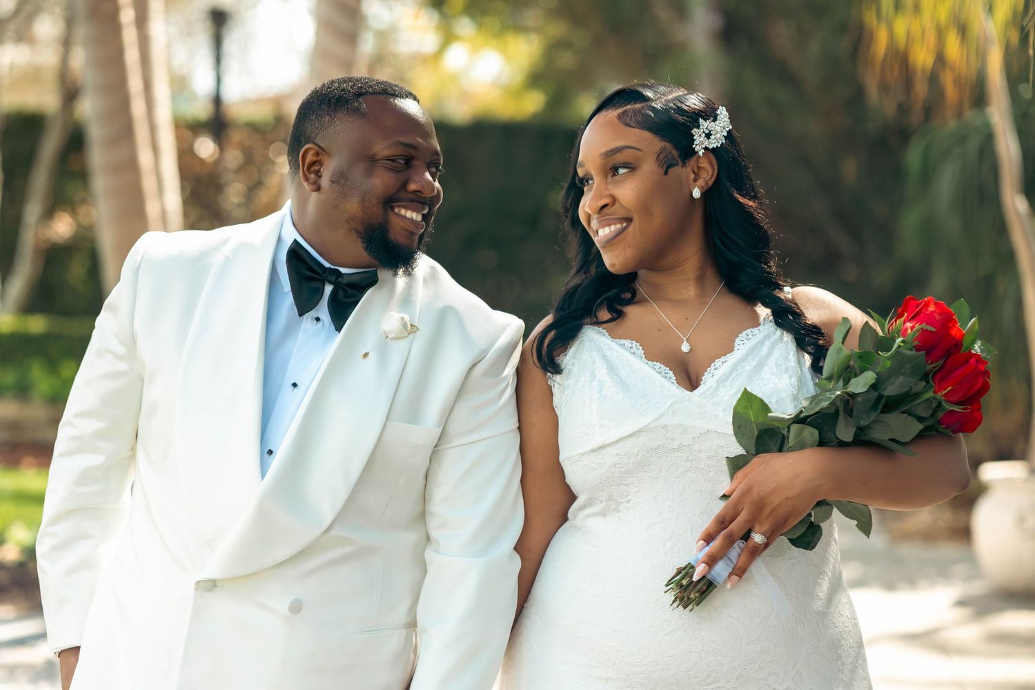Happy newlywed couple in white wedding attire, bride holding red roses bouquet, smiling outdoors on their wedding day.