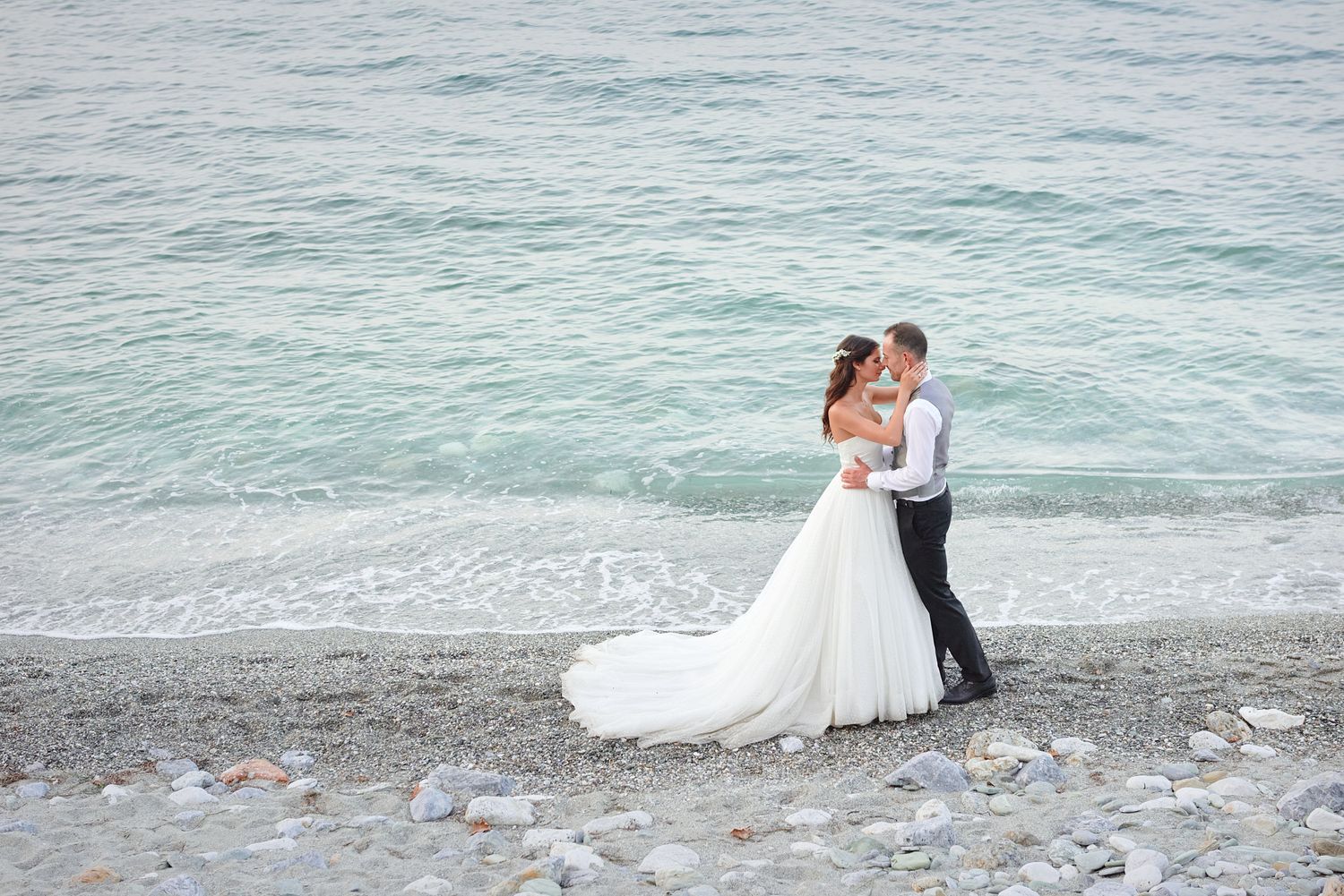 Distant full-length view of a newly married couple embracing on a rocky Greek beach.