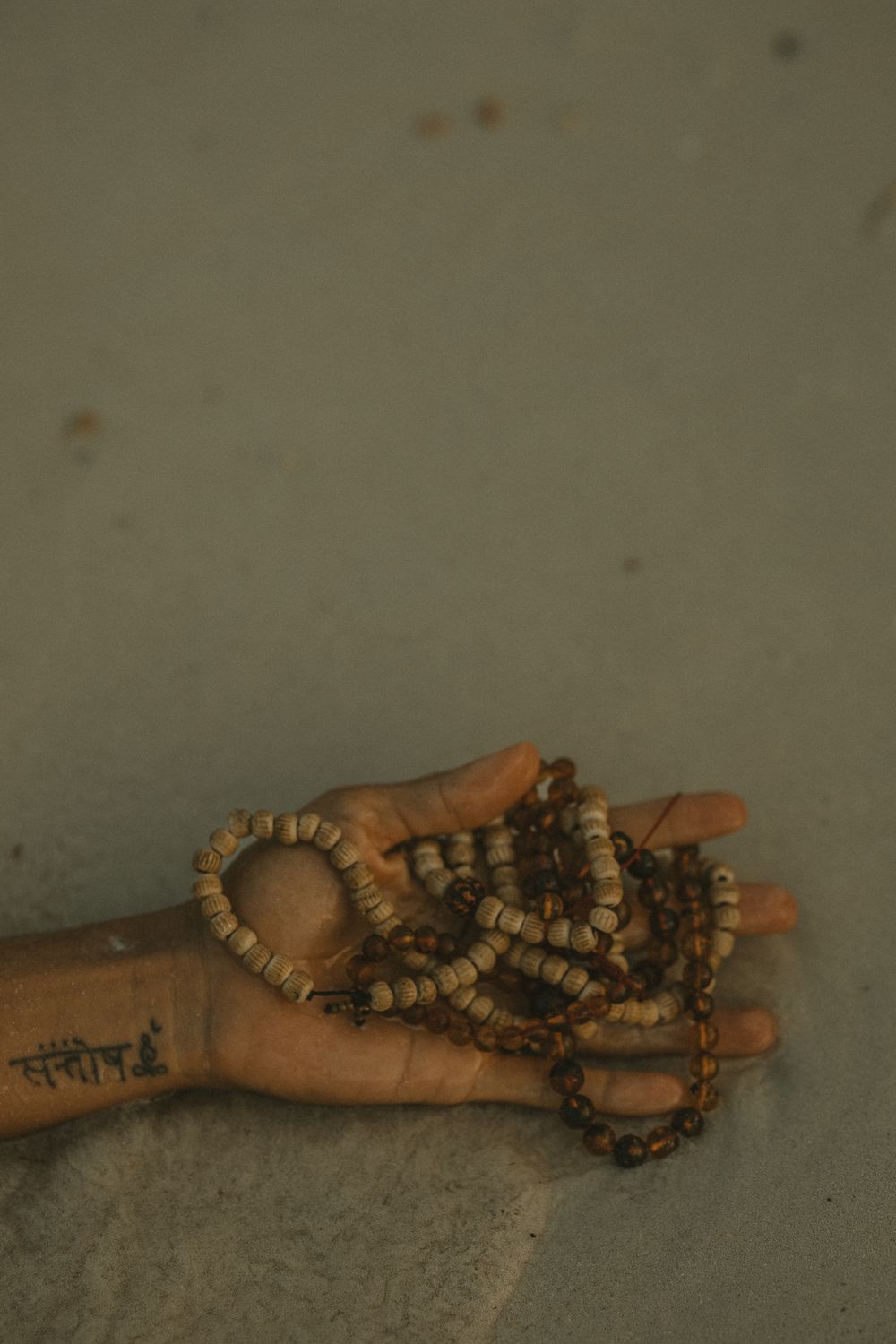 Hand holding wooden prayer mala beads with a Sanskrit tattoo on the wrist against grey background.
