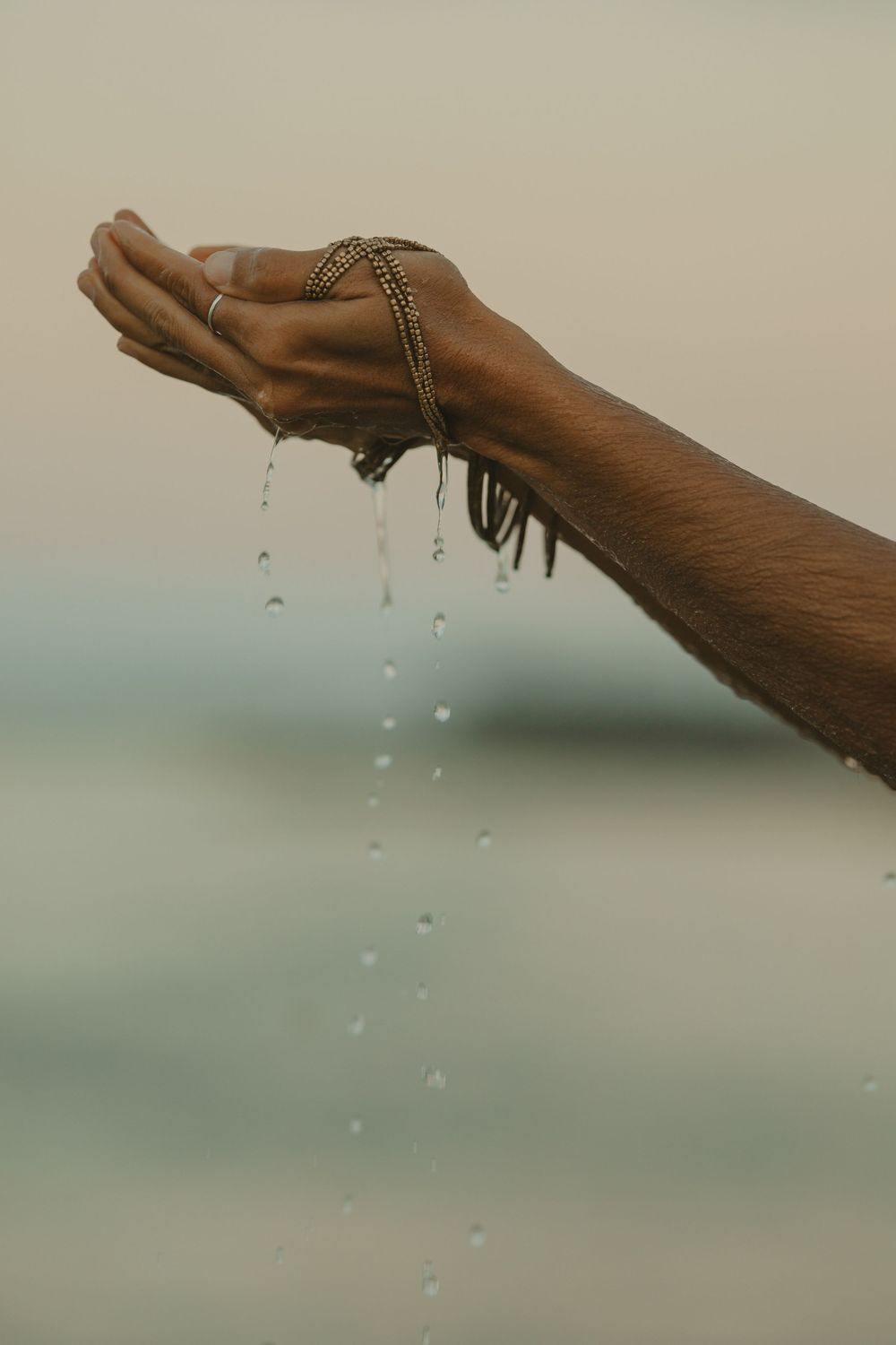 Water droplets falling from a raised dark-skinned hand wearing a gold bracelet against misty background.