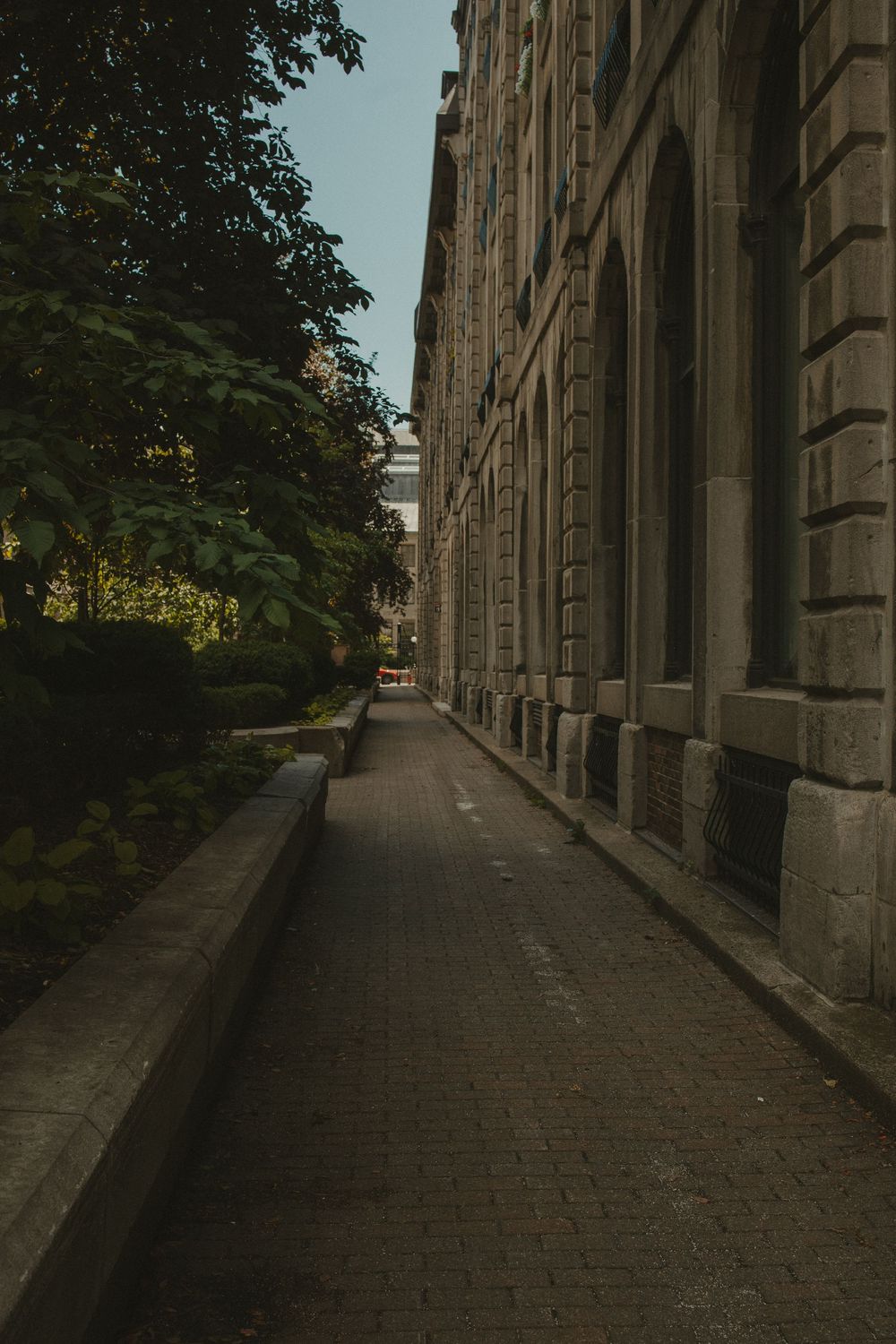 Narrow brick pathway alongside a historic stone building with trees and greenery on the opposite side.