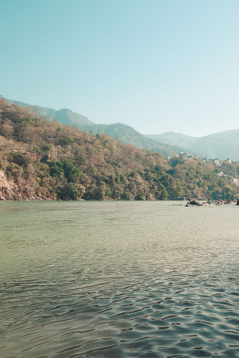 Calm river with gentle ripples reflecting mountains covered in lush trees under a clear blue sky.