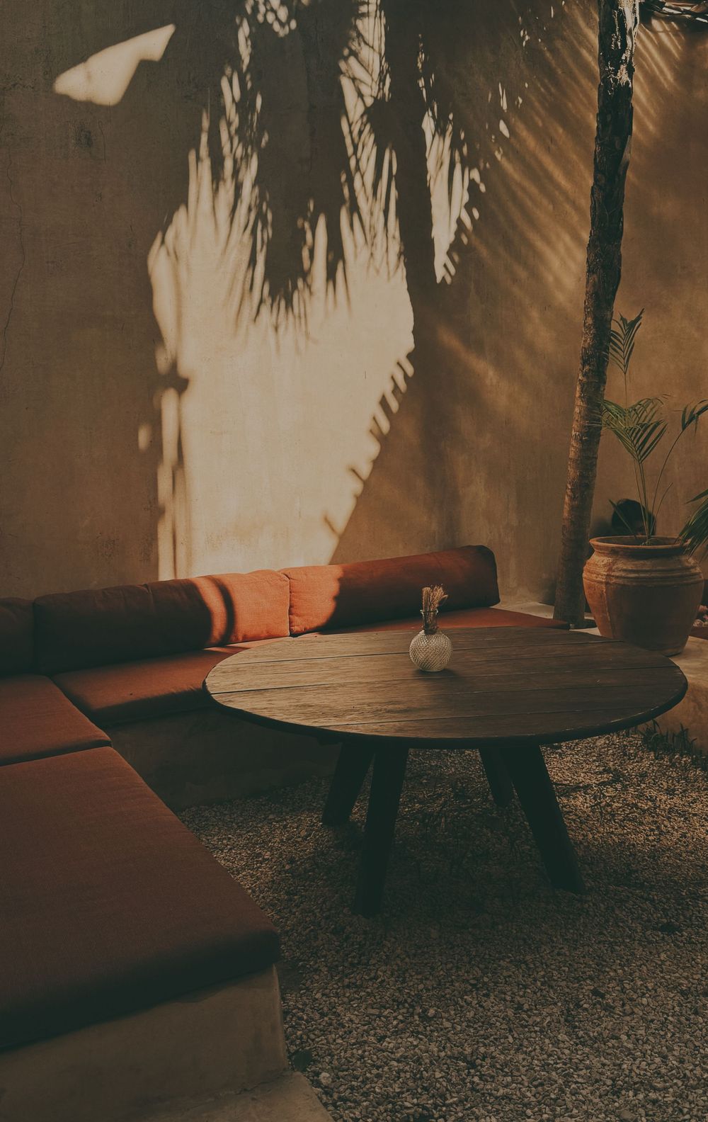 Warm sunlight casts palm shadows on a rustic lounge with orange cushions and round wooden table.
