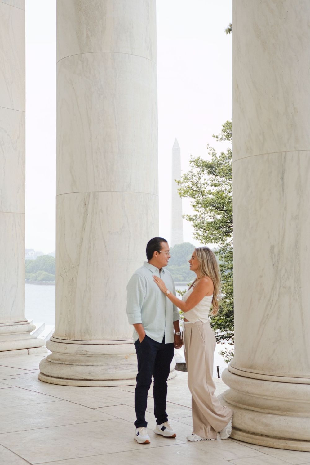 Couple shares romantic moment between marble columns at Jefferson Memorial with Washington Monument visible in background.
