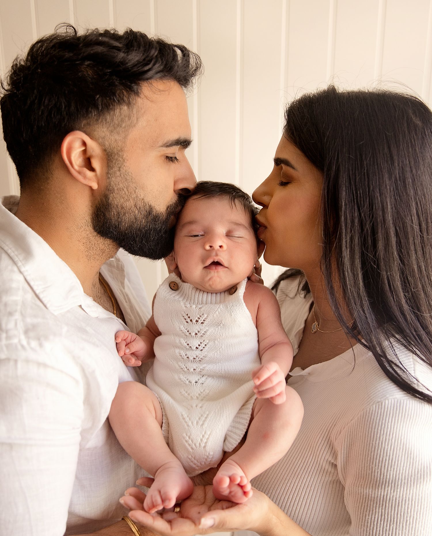 Two parents kissing their newborn baby on the cheeks, holding the infant in a softly lit family portrait. During a newborn session London