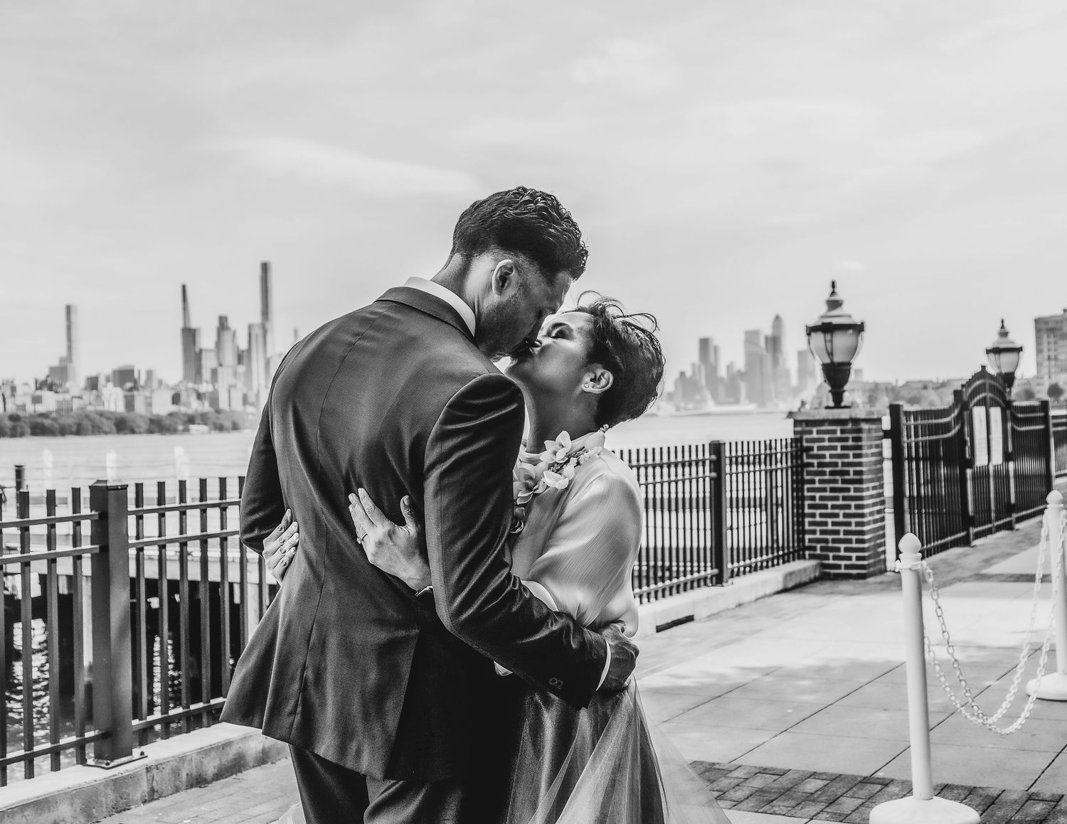 Black and white photo of a couple sharing a romantic kiss on a waterfront promenade with a city skyline backdrop.