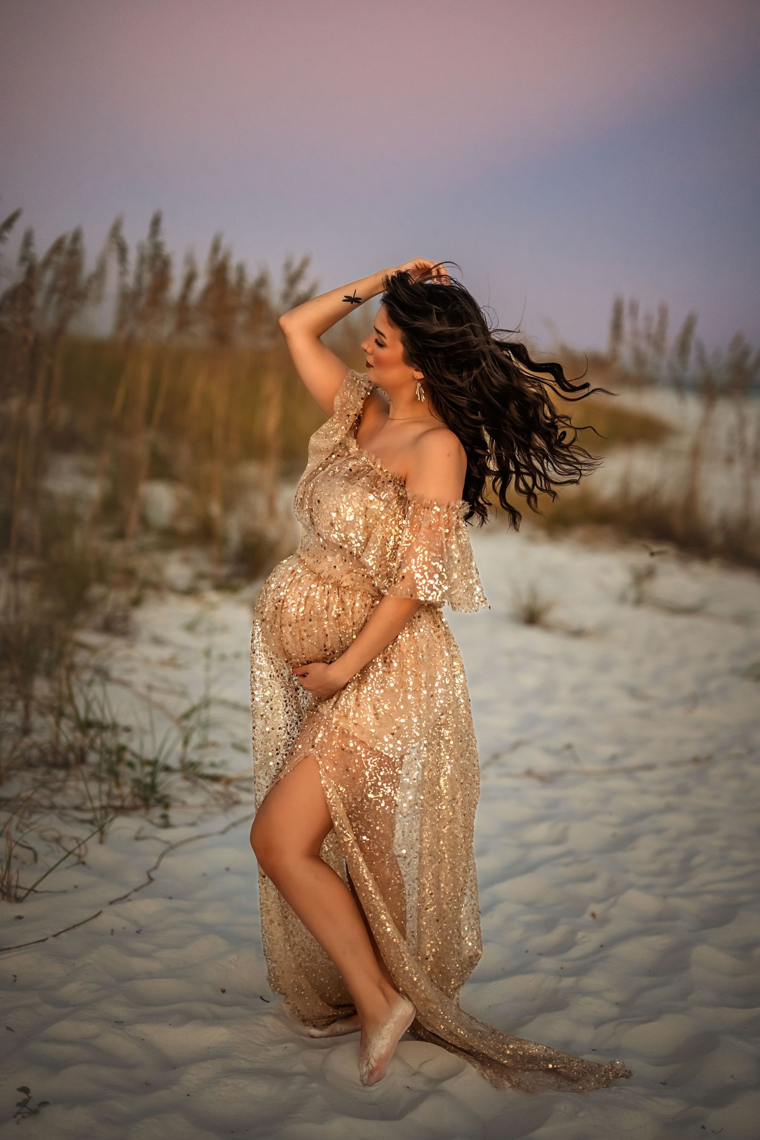 Pregnant woman in gold sequin off-shoulder gown poses on white sand beach at sunset with wind-blown hair.
