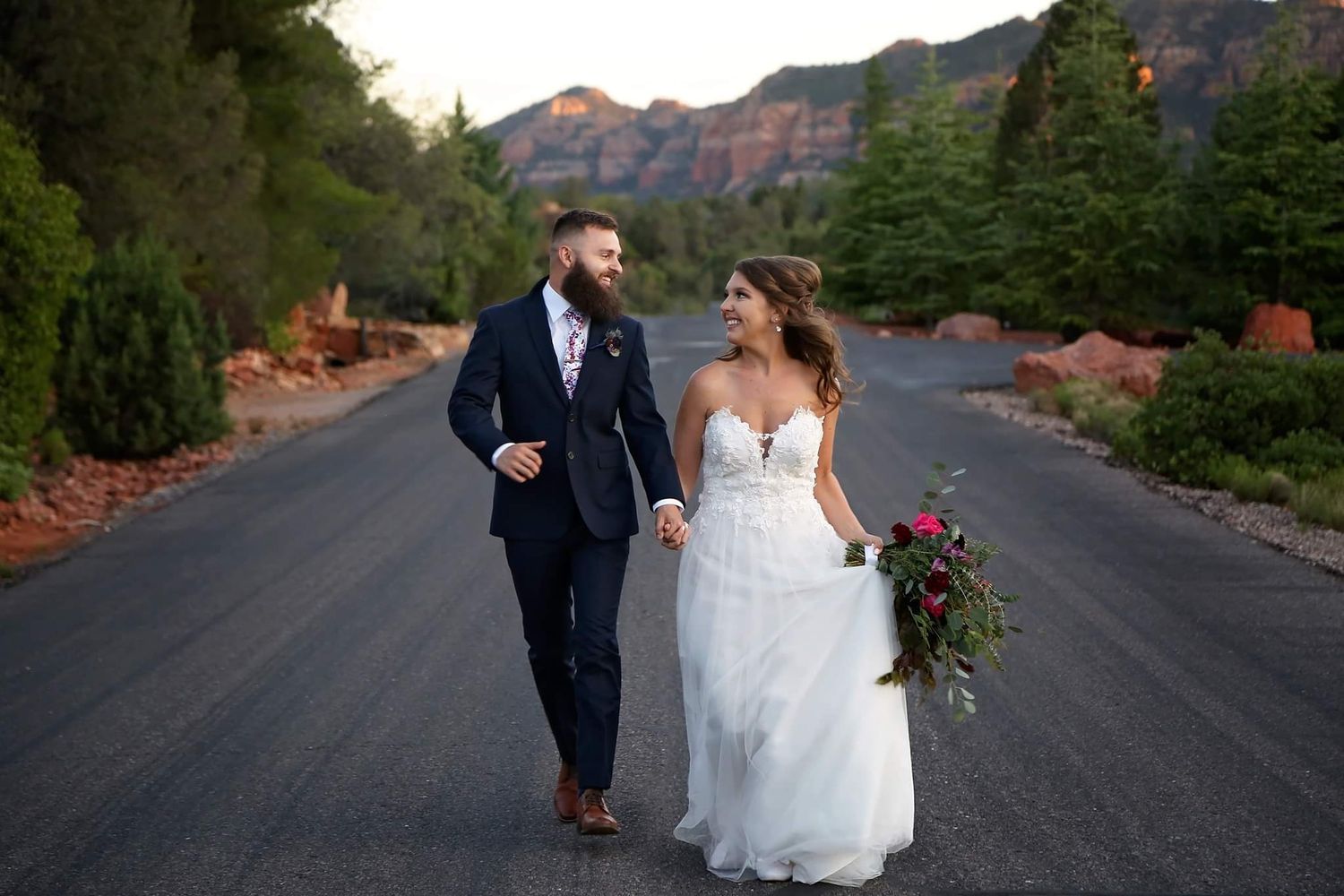 A bride in a flowing white gown holds hands with her groom while walking down a road lined with red rock and greenery.