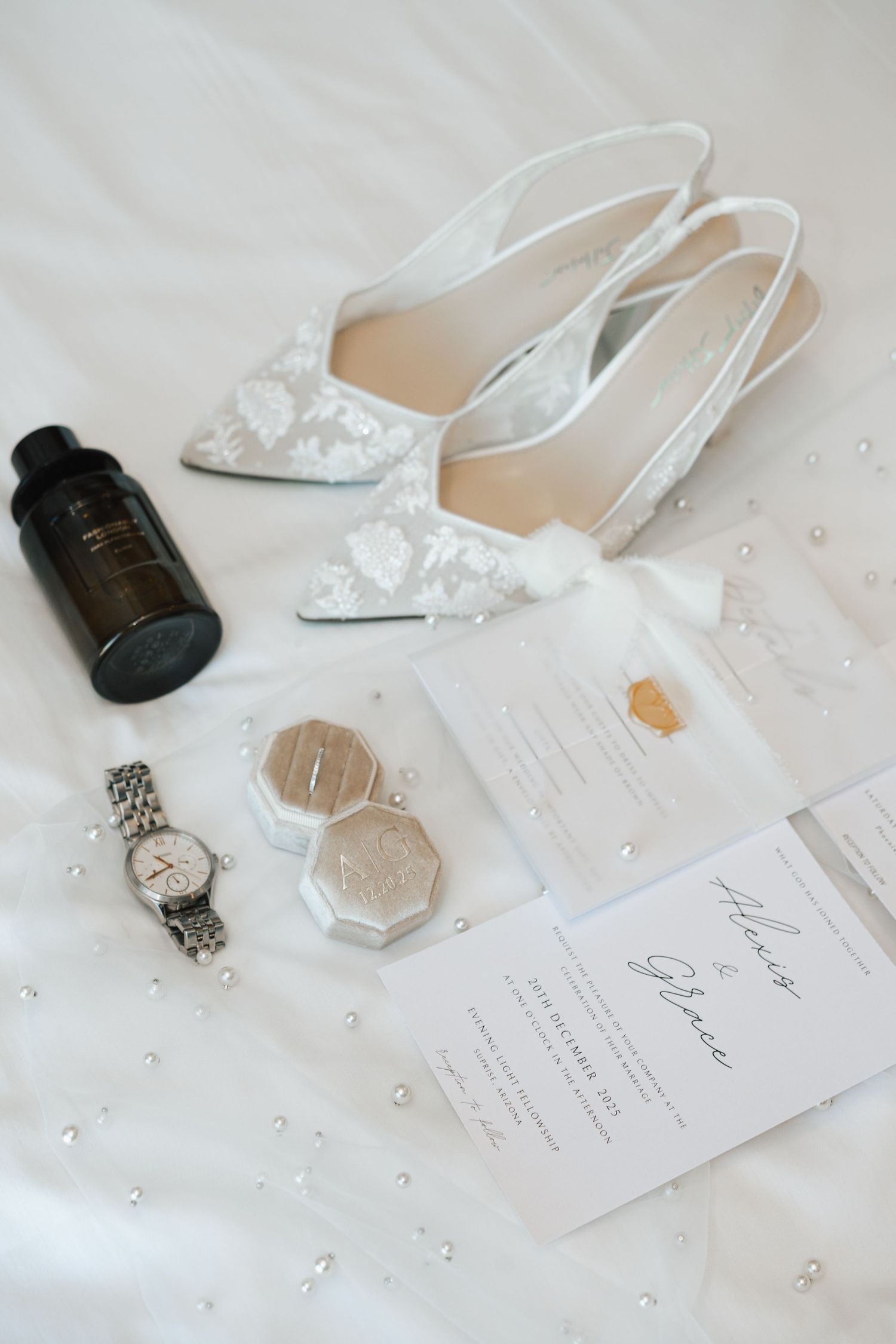 Elegant bridal flat lay with lace slingback heels, wedding rings, watch, perfume, and invitation suite on white fabric.