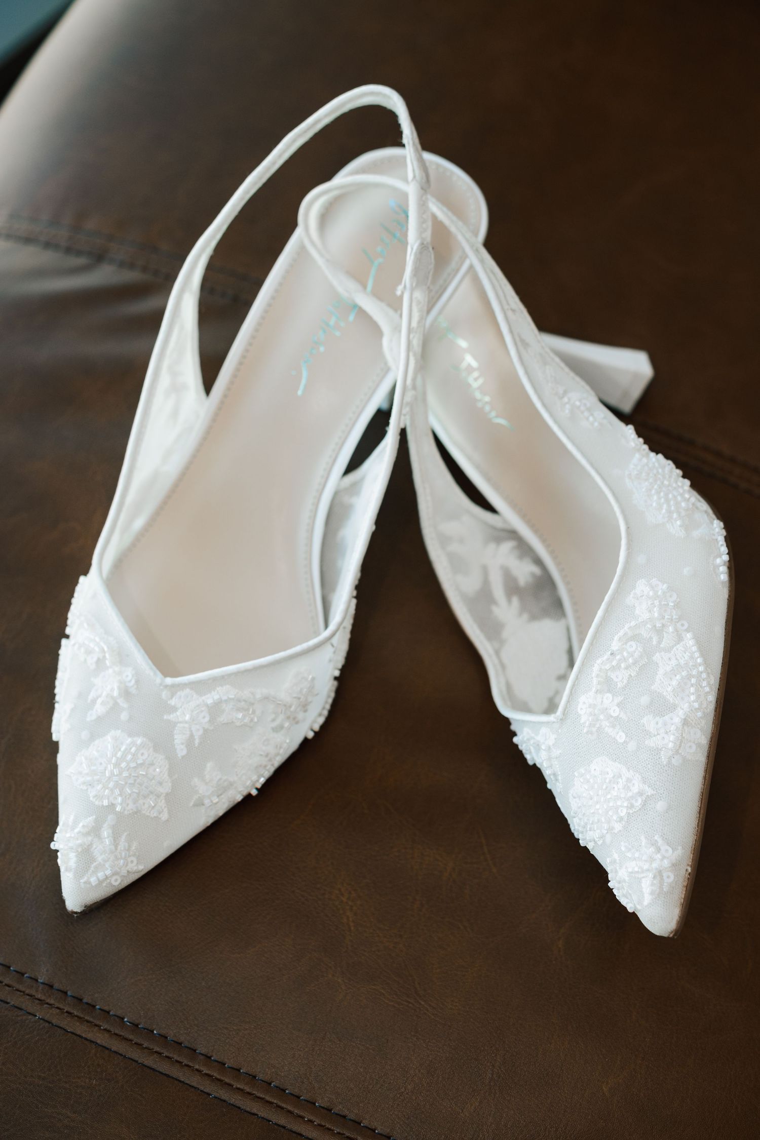 White lace slingback bridal heels with pointed toe and floral embroidery displayed on brown leather surface.