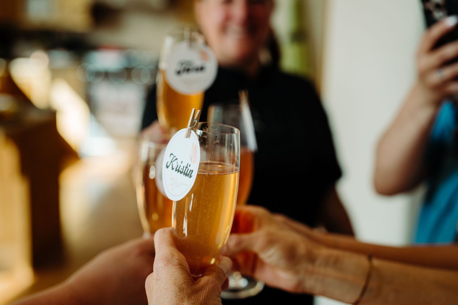 Hands clinking champagne flutes in a celebratory toast, with a decorative tag on one glass at a bar.