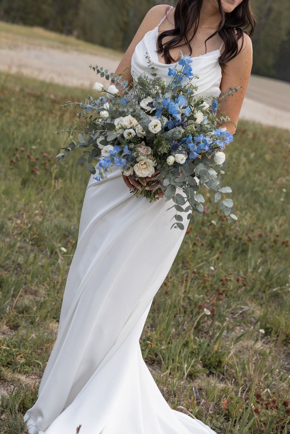 Bride in white satin gown holding lush blue and white wedding bouquet with eucalyptus in outdoor field setting.