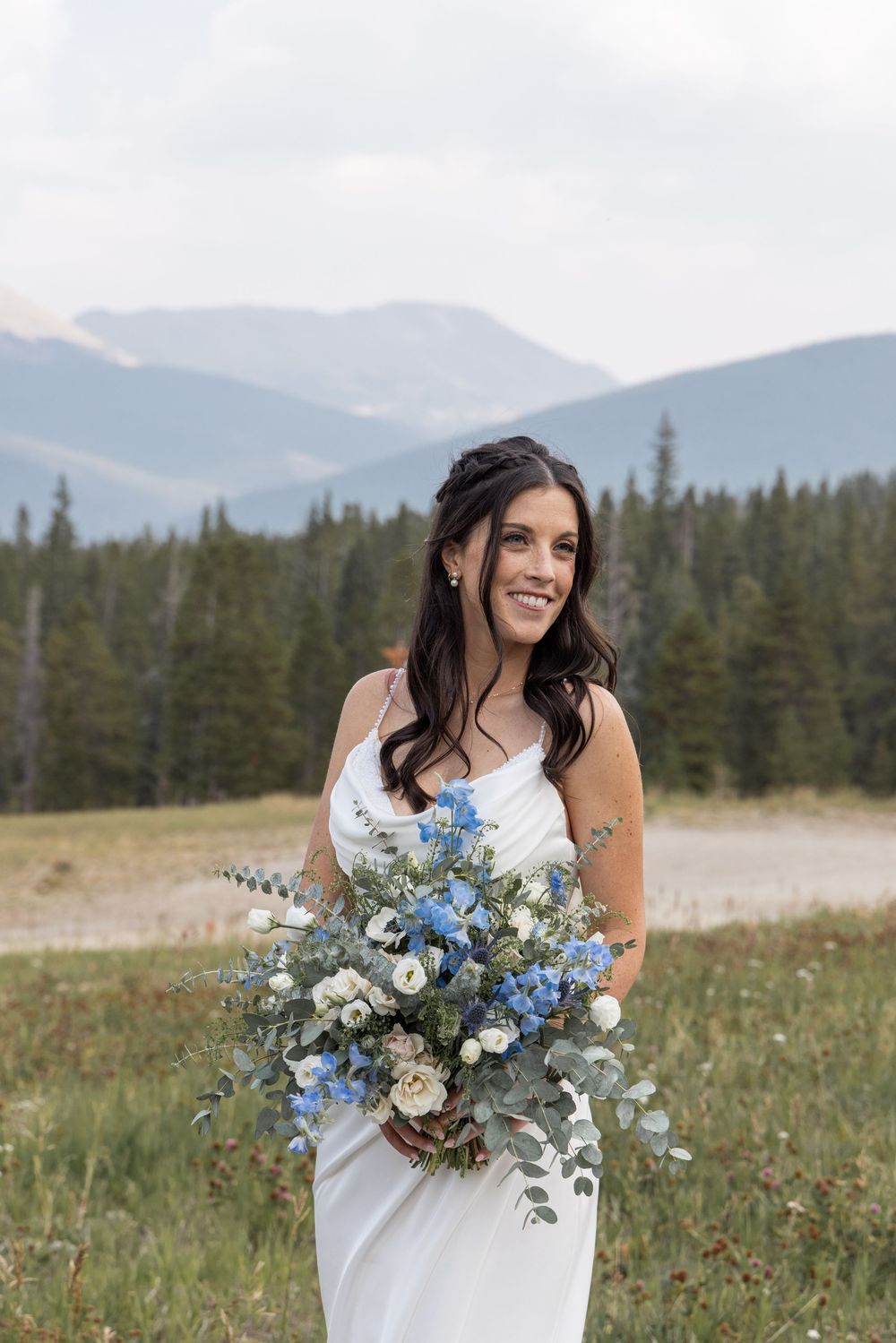 Smiling bride in white gown holding blue and white bouquet with mountain forest backdrop.